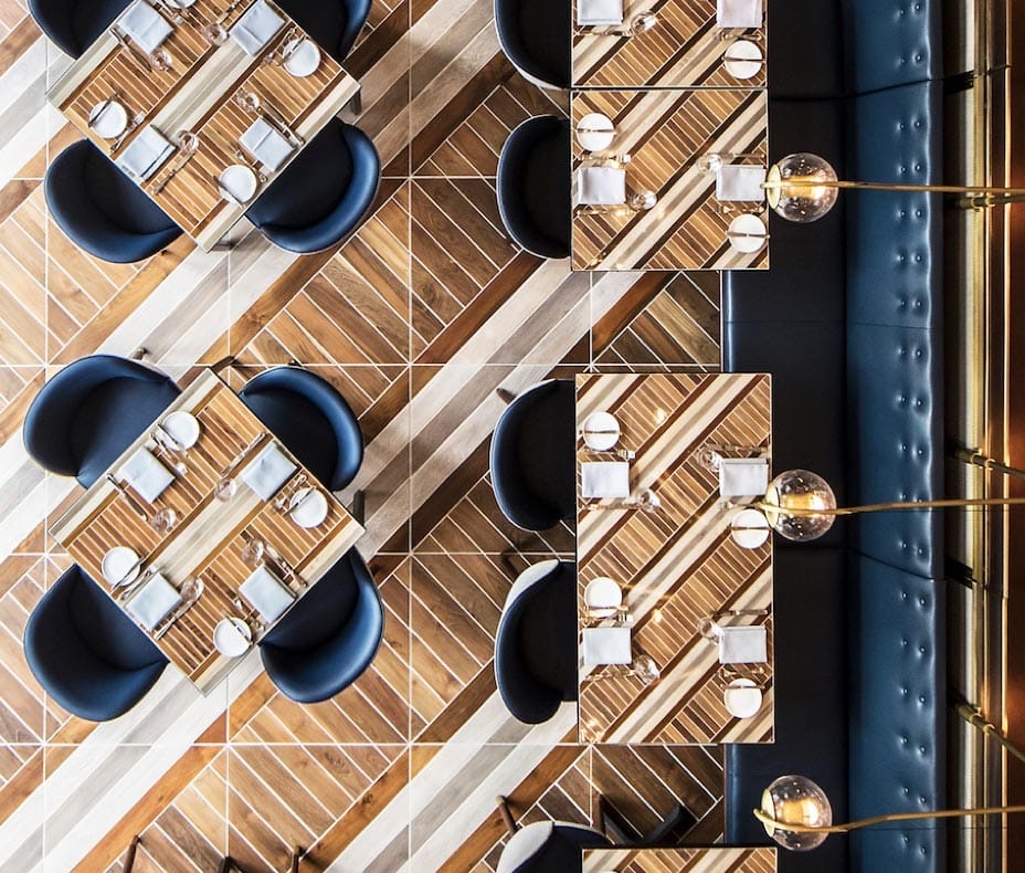 Overhead view of a restaurant with a diagonal teak and white-stripe floor, navy chairs, and brass globe lamps